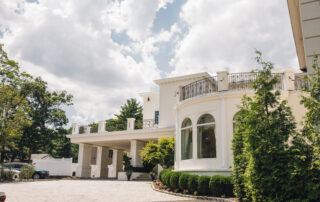 Large white house with circular windows, a balcony, columns, and a driveway under a partly cloudy sky; landscaped bushes and trees line the front, evoking the elegance of a Tides Estate.