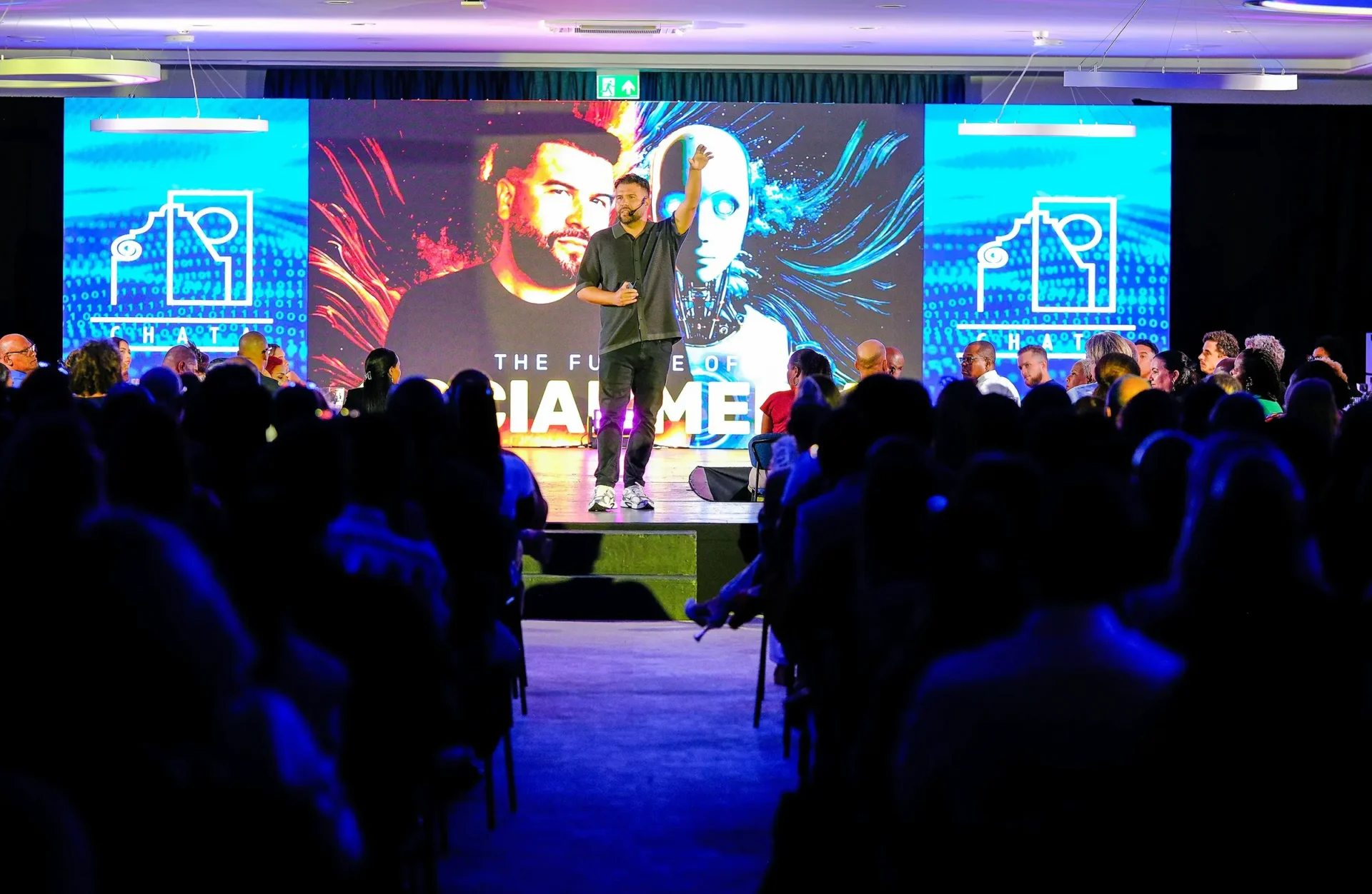 Speaker on a stage with LED video walls and purple lighting in front of a full corporate audience